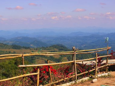 Manzara bir bambu çiti ve parlak kırmızı çiçekler içerir, arka planda hafif bir sisle örtülü katmanlı dağlarla..