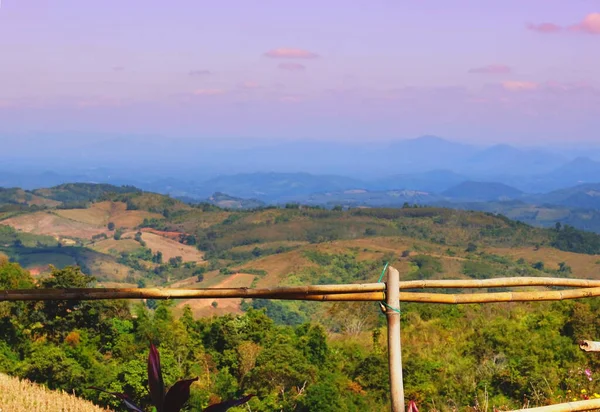 A landscape of rolling hills and mountains in the distance, with a bamboo fence in the distance. The area is filled with lush greenery.