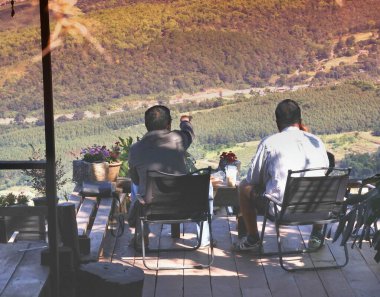 İki adam ahşap bir güvertede, güneşli bir günde vadiyi ve yemyeşil ormanı kuşbakışı seyrediyor..