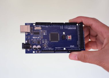 Mavi bir Arduino Mega 2560 Rev3 mikro kontrol panosunu beyaz bir arkaplanda tutan bir elin yakın plan görüntüsü..