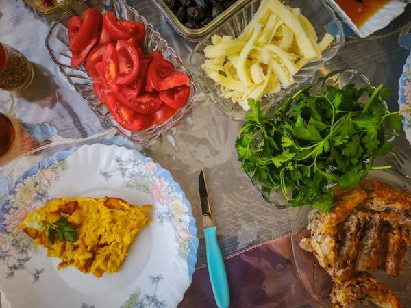 Türk kahvaltı masası. İnsanların elleri hamur işlerini, sebzeleri, yeşillikleri, zeytinleri, peynirleri, kızarmış yumurtaları, baharatları, reçelleri, balı, bakır demlikte çayı ve lale bardaklarını, geniş kompozisyonu alıyor.