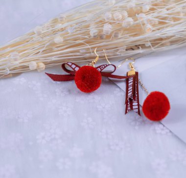 Kırmızı ponpon küpeler, şekli tüylü bir toptur. Küpelerin yanında karton ve buket numuneleri gibi dekoratif nesneler de var. Bu eşyalar tülün üzerine yerleştiriliyor. Küpeler çok zarif ve şık görünüyor..