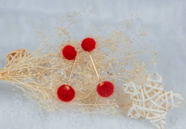 Kırmızı ponpon küpeler iki kabarık topa benzer. Küpelerin yanında rattan ürünleri ve buket örnekleri var. Bu eşyalar tülün üzerine yerleştiriliyor. Küpeler çok güzel ve şık görünüyor..