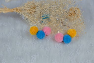 Üç kürklü top şeklinde renkli ponpon küpeler. Küpeler, rattan ürünleri, çiçek numuneleri ve diğer süslemelerin yanındadır.