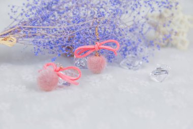 Pembe ponpon küpeler, ayrıca bir de yayı var. Küpelerin yanında mavi buket örnekleri, çeşitli şekillerde rattan ürünleri ve diğer dekoratif nesneler var. Bu eşyalar tülün üzerine yerleştiriliyor. Bu kombinasyonlar zarif ve şık görünüyor..