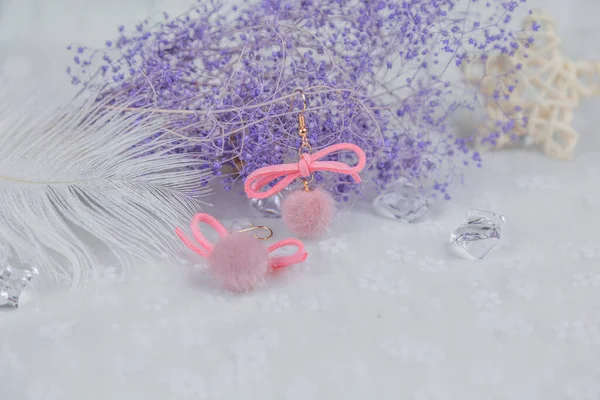 Pembe ponpon küpeler, ayrıca bir de yayı var. Küpelerin yanında mavi buket örnekleri, çeşitli şekillerde rattan ürünleri ve diğer dekoratif nesneler var. Bu eşyalar tülün üzerine yerleştiriliyor. Bu kombinasyonlar zarif ve şık görünüyor..