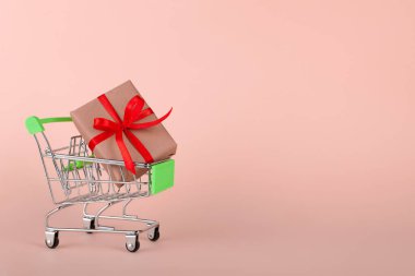 Pembe arka planda mini market arabasında kırmızı kurdeleli el işi hediye kutusu.