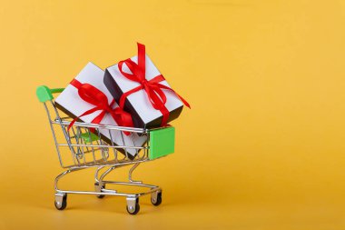 Kırmızı kalp, içinde kırmızı kurdele olan hediye kutusu renkli arka planda mini market arabasının içinde.