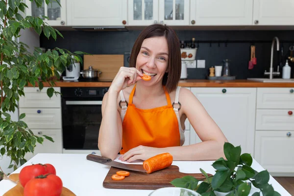 Joven hermosa mujer sentada en el fondo de la cocina mientras corta verduras Fotos De Stock Sin Royalties Gratis