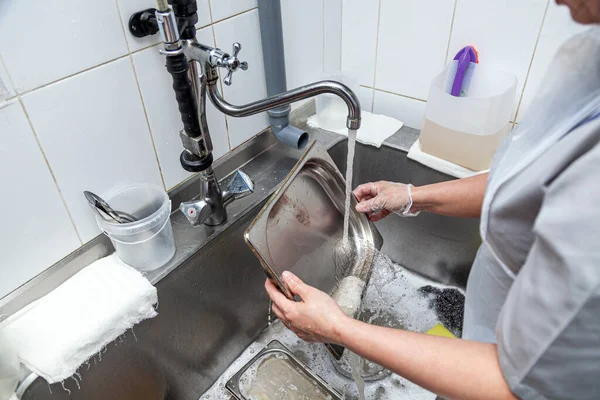 Lavavajillas en uniforme lava plancha con espuma y esponja bajo el agua del grifo —  Fotos de Stock