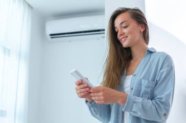Genç, mutlu, gülümseyen kadın uzaktan kumandayı kullanarak klimanın sıcaklığını ayarlıyor..