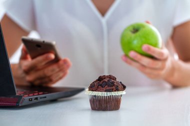 İşte yemek yemek, ofiste öğle yemeği molası. İş kadını telefon ve dizüstü bilgisayar başında çalışırken elmalı ve çikolatalı kek yiyor. 