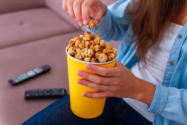 女性座っています上のソファと食べるカリカリポップコーンの間にテレビを見ますホーム — ストック写真