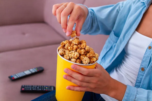 女性座っています上のソファと食べるカリカリポップコーンのために軽食のためにテレビを見ますホーム — ストック写真