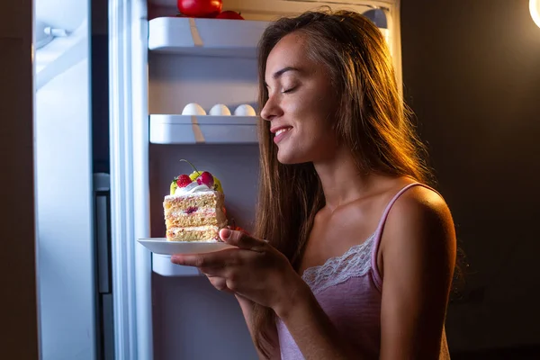 Hungry woman in pajamas eats sweet cake at night near refrigerator. Stop diet and gain extra pounds due to carbs food and unhealthy eating