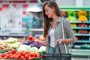 Kadın süpermarketin sebze bölümünden taze olgun domates seçiyor. Müşteri marketten yiyecek alıyor.