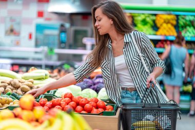 Alışveriş sepeti olan kadın sebze bölümünden taze kırmızı domates seçiyor. Müşteri marketten yiyecek alıyor.