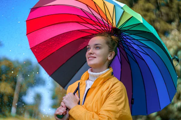 Happy woman in a yellow jacket with multi-colored rainbow umbrella during the rain outdoors in rainy weather