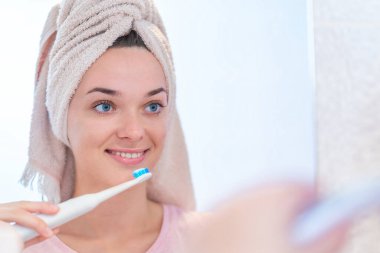 Mutlu ve sağlıklı bir kadın sabahın erken saatlerinde evdeki banyoda ultrasonik elektrikli diş fırçası kullanarak dişlerini fırçalıyor. Ağız temizliği, diş ve diş sağlığı, sağlıklı dişler. Günlük hayat ve rutin