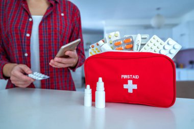 Woman using smartphone for reading and examining medicine instruction on the internet before take pills. First aid kit on table at home