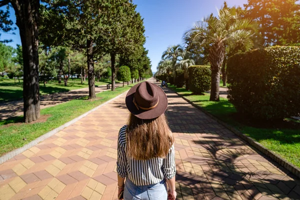 ヤシの木と木の間の公園の路地に沿って晴れた日に午後に歩く帽子のブルネットの女性 — ストック写真