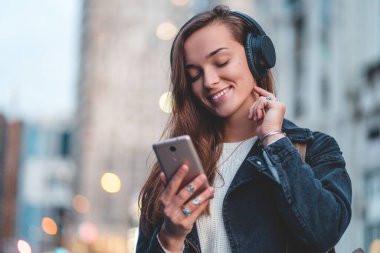 Young happy stylish trendy casual hipster woman changes songs and tracks on smartphone during listening to music on a wireless headphone while walking around the city. Music lover enjoying music clipart