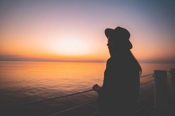 Silhouette of one lonely traveler woman in a hat during standing on sea pier and looking into the distance at sunset