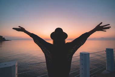 Günbatımında deniz rıhtımında açık kolları olan, şapkalı, yalnız, özgür bir gezginin silueti. Mutlu özgürlük kavramı ve hayatın tadını çıkarmak