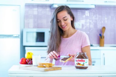 Batumi, Georgia - 11 Kasım 2019. Mutlu, çekici, genç, neşeli kadın evde tatlı olarak tost ekmeğine fındıklı çikolata ezmesi sürüyor. 