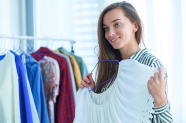 Mağazada kıyafet alışverişi yaparken modern modaya uygun moda kadın kıyafetleri deneyen ve giyecek kıyafet seçen mutlu, genç esmer kadının portresi. 