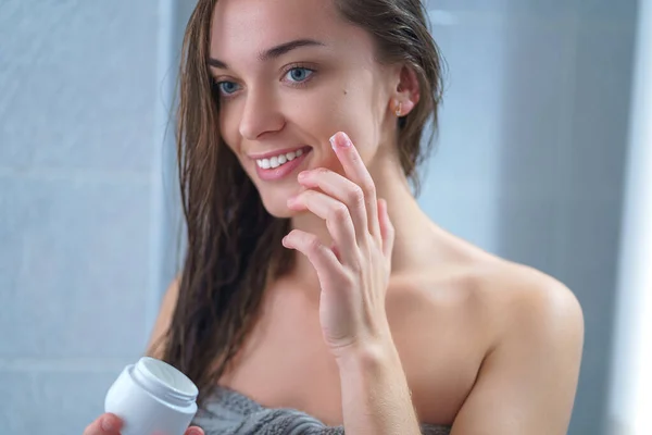 タオルで幸せな健康的な魅力的な若い女性は 自宅でシャワーした後 バスルームできれいな顔に保湿クリームを適用します 肌のケアと水分補給 女性のケア — ストック写真
