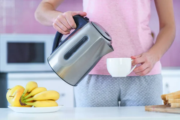 Woman pouring hot water from electric kettle for brewing tea for tea time in the kitchen at home