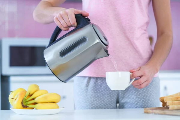 Woman pouring hot water from electric kettle for brewing tea for tea break in the kitchen at home