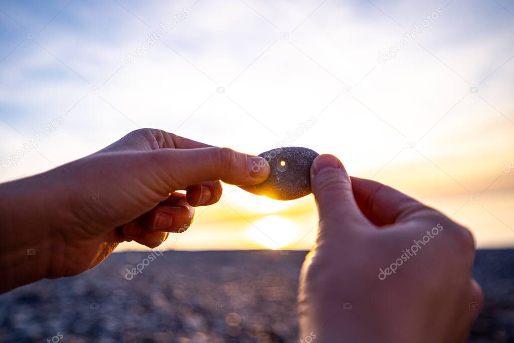 Person holds a stone with a hole at sunset. Find a mascot chicken god and make a wish