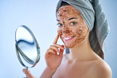 Yüzü doğal havluyla gülümseyen mutlu kadın duştan sonra maskesini yuvarlak küçük bir aynayla fırçalayıp spa günü ve evde cilt bakımı rutini yapıyor.