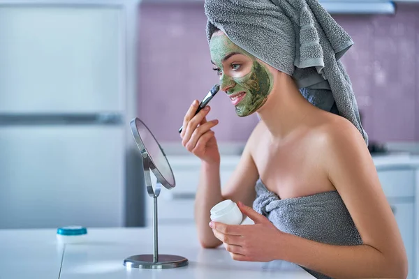 Young healthy woman in bath towel applying cleansing face clay mask after shower during spa day at home using brush and small round table mirror. Face skin care
