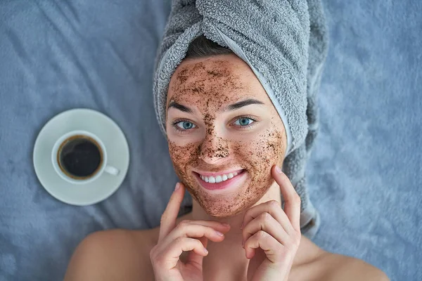 Portrait of smiling healthy happy female in bath towel with natural face coffee scrub during spa day and skin care routine at home