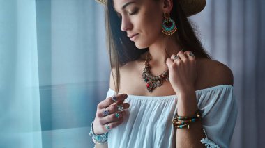 Zarif esmer boho şık kadın beyaz bluz ve büyük küpeli hasır şapka takıyor, bilezikler, altın kolye ve gümüş yüzükler. Mücevher detaylarıyla süslü moda çingene bohem kıyafeti. 