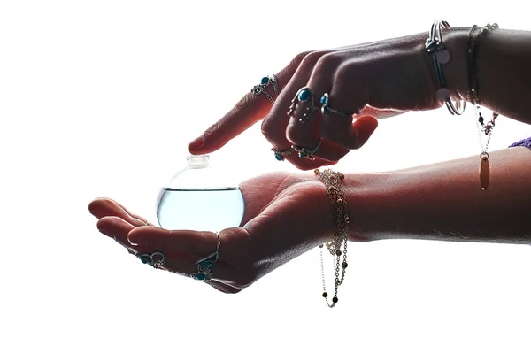 Crystal elixir potion bottle for love spelling, witchcraft and divination in perfumer woman hands wearing silver rings with turquoise stone and bracelets on a white background. Alchemy and perfumery