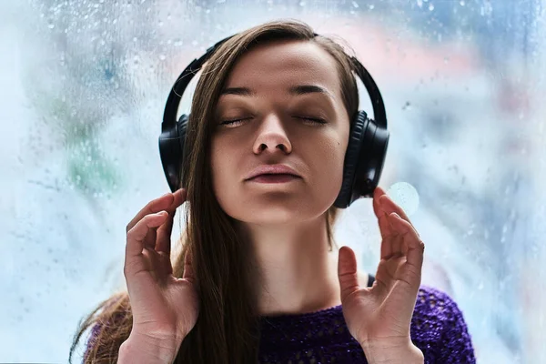 Woman music lover with closed eyes in wireless headphones enjoys and listens to soothing calming relaxing sound during rainy autumn weather