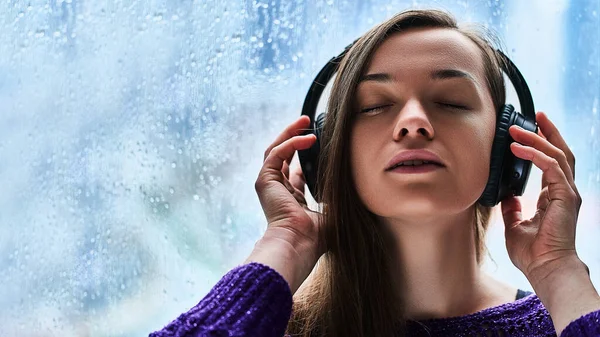 Woman music lover with closed eyes in wireless headphones enjoys and listens to soothing calming relaxing music during rainy autumn weather Copy space