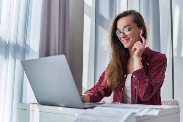 家庭で働くリモートオンラインブログのためのコンピュータを使用してワイヤレスヘッドフォンやメガネで幸せなヒップスターフリーランスの女性 — ストック写真