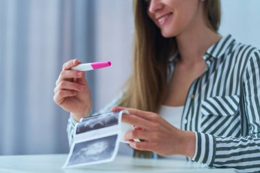 Happy smiling woman looking at a positive pregnancy test result and holding ultrasound picture. Planned and long-awaited pregnancy clipart