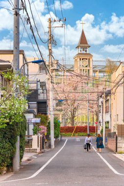 Tokyo, Japonya - Nisan 07 2021 Seigakuin İlköğretim Okulu 'nun çan kulesinin altında bir Hıristiyan haçı tarafından gözden kaçırılmış ve saat ve karilyon çanları ile süslenmiş kiraz ağaçlarıyla çevrili sokak..