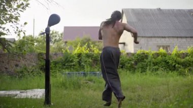Uzun siyah saçlı, güzel gövdeli, bahçede karate yapan, tekme pratiği yapan atletik bir adam.