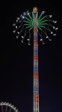 Gece aydınlatmalı salıncak, dönen tepe örtüsüyle, sallanan sandalye ve tellerle parlayan dev parlayan eğlence kulesi, parlayan teller ve dönen tepe örtüsüyle parlayan gökyüzü yüksek eğlence gezisi. 4k