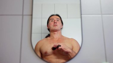 Bir adam banyoda aynanın önünde göğsünü elektrikli jiletle tıraş ediyor. Tişörtsüz bir adam banyo aynasında kendine güvenini ve özgüvenini sergiliyor..
