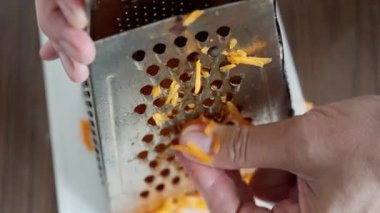 Yetişkin parmakları peyniri konteynırın üstünde rendeliyor, çedar peyniri tek tek rendeliyor, insan çedar peynirini ince ince rendeliyor mutfaktaki metal peynir rendesinin üstünde rendeliyor.