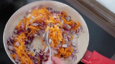 Tavada doğranmış havuç ve kırmızı soğan, sobanın üzerinde kaşıkla karıştırılan ellerin yakın çekim görüntüsü, sıcak ışık, canlı turuncu ve mor sebzeler ev aşçılığı yaparken cızırdıyor.