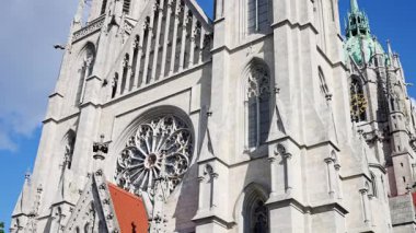 Mavi gökyüzünün altındaki gotik katedral cephesi, dekoratif taş işçiliği ve dev gül penceresi, yükselen kule ve kemerli girişin üstündeki kırmızı kiremitli kubbe, güneş ışığı dramatik gölgeler bırakıyor.